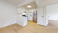 This 540 sq ft, single-floor apartment features 2 bedrooms and 1 bathroom with a clean, modern aesthetic. Light hardwood flooring runs throughout, complementing crisp white walls and trim. The open kitchen boasts white shaker cabinets, marble-style countertops, stainless steel appliances, and a peninsula. The living area offers natural light via large windows with blinds. The primary bedroom measures 150 sq ft with a dedicated window. The bathroom features white subway tile wainscoting, a white vanity, and black hardware accents. A hallway with closet access connects all rooms efficiently.