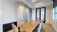 This single-floor professional space is designed to foster productivity and collaboration in a polished, modern environment. The centerpiece of the room is a rectangular wooden conference table surrounded by ergonomic high-back chairs upholstered in light grey, offering comfortable seating for a full team. The room's design is defined by its clean, neutral color palette — soft grey and muted blue wall panels lend a calm, focused atmosphere, while crisp white ceilings amplify the sense of openness. Overhead, a combination of recessed linear LED lighting and directional track lights ensures even, well-distributed illumination throughout the space. Built-in floor-to-ceiling cabinetry along one wall provides ample storage while maintaining the room's streamlined aesthetic. A glass partition wall with a frosted privacy film and dark framing elegantly separates the conference room from the adjacent area, allowing natural light to filter through while maintaining a degree of visual privacy. The carpeted flooring adds a subtle warmth and helps with acoustics, making the room well-suited for focused meetings and discussions. Overall, the space exudes a refined, corporate sophistication ideal for professional gatherings.