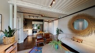This single-floor shipping container residence showcases a striking industrial-chic aesthetic, where the structure's origins are celebrated through exposed corrugated metal ceilings painted white, visible black pipe conduits, and track lighting throughout. The open-plan living and kitchen area forms the heart of the home, where rich dark hardwood floors ground the space and connect the functional zones seamlessly. The kitchen is outfitted with stainless steel Whirlpool appliances—including a range, built-in microwave, dishwasher, and full-size refrigerator—complemented by white shaker cabinetry, clean white countertops, and open metal shelving for additional storage. A dining area with a wood table flows naturally into the living space, which features a light sofa, leather accent chairs, and lush indoor plants that soften the industrial elements. Floor-to-ceiling glass doors open onto a private balcony with outdoor views, flooding the area with natural light. The bedroom offers a serene retreat with neutral tones, a large window, and the signature corrugated ceiling detail. A clean, well-appointed bathroom with a white vanity and large mirror completes the layout. The overall ambiance balances raw industrial character with warm, curated décor.