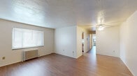 This single-floor residence offers a practical and comfortable layout with consistent hardwood-style flooring running throughout the living areas, bedroom, and kitchen. The open living room is generously proportioned, featuring a textured ceiling, a classic cast-iron radiator for heating, and a large window fitted with horizontal blinds that allows natural light to fill the space. A ceiling fan provides additional comfort and air circulation. The living area connects seamlessly to a hallway that leads to the remaining rooms.

The galley-style kitchen is well-appointed with warm medium-toned wood cabinetry on both sides, light granite-style countertops, and white appliances including a refrigerator, electric range, and sink. A window above the radiator brings in natural light and offers a view of the surrounding exterior.

The bedroom is a well-sized room benefiting from two windows that provide ample daylight and views to the outside. A ceiling fan with integrated lighting adds to the room's functionality, and a cast-iron radiator ensures consistent warmth.

The bathroom features classic white tile with teal accent detailing along the walls, a mosaic tile floor, a full shower-tub combination with curtain rod, and a small frosted window for natural light and privacy. The space is clean and neatly arranged. Overall, the residence presents a functional, straightforward layout with consistent finishes throughout.