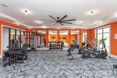 Located at 1035 Old Peachtree Rd NW in Suwanee, Georgia, this well-appointed fitness center delivers a vibrant and energizing workout environment across a single open floor. The space is immediately distinguished by its bold orange accent walls, which contrast sharply with crisp white ceilings and trim, creating a high-energy atmosphere designed to motivate. A bold geometric-patterned carpet covers the main exercise floor, while a dedicated cycling and free weights studio features smooth hardwood-style flooring for a clean, functional transition between zones. The main workout area is generously sized and equipped with treadmills, elliptical trainers, rowing machines, and strength training equipment, all arranged to allow ample movement between stations. A large dumbbell rack and additional selectorized strength machines round out the offerings. Large floor-to-ceiling windows line multiple walls, flooding the space with natural light and offering views of the surrounding outdoor pool area and landscaping. Ceiling fans provide additional airflow throughout. The cycling studio houses stationary bikes alongside a dumbbell rack and bench, with three wall-mounted flat-screen televisions to keep users entertained during workouts. Recessed and flush-mount lighting ensures the space remains well-lit at all hours, and full-length mirrors in the main area enhance the sense of openness.