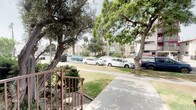 This single-floor residential building presents a classic Southern California architectural style, characterized by a textured stucco exterior finished in a warm yellow tone with Spanish-influenced terra cotta roof tile accents visible along the roofline. The building's façade features a decorative upper-level balcony with lattice detailing, adding visual interest to the street-facing elevation.

Entry to the building is secured through ornate wrought iron security gates with scrollwork detailing, complemented by a wall-mounted intercom system for controlled access. The brick accent walls flanking the entryway add a layer of architectural texture and durability to the building's approach.

A concrete pathway leads from the street to the main entrance steps, which are flanked by mature, established landscaping including shrubs, tropical plants, and potted greenery that lend a welcoming, verdant character to the exterior. Large, mature trees line the property and the surrounding street, providing ample shade and a sense of established greenery throughout the neighborhood.

The surrounding streetscape is residential in character, with similar multi-unit buildings nearby, wide sidewalks, and well-maintained lawns. The property benefits from a peaceful street-level outlook framed by the canopy of mature trees, creating an inviting and serene environment. The building's gated perimeter and intercom system provide an added layer of security and privacy for its residents.