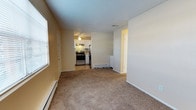 This single-floor space offers a practical and comfortable layout that connects the main living area to a well-equipped kitchen. The living room features wall-to-wall carpet in a neutral tan tone, providing a warm and understated foundation for the space. Natural light filters in through a window fitted with blinds, brightening the room alongside baseboard heating units positioned along the walls, ensuring year-round comfort. The textured popcorn ceiling adds a classic residential character throughout.

The kitchen is visible from the living area and is outfitted with stainless steel appliances, including a gas range and refrigerator. A patterned tile backsplash introduces a subtle decorative element, while under-cabinet lighting and a ceiling fan with a light fixture add both function and ambiance. The kitchen floor transitions to a hard surface, distinguishing it from the carpeted living space.

Two doors are visible — one appearing to lead to a closet and another to an additional room or hallway — suggesting a thoughtful, efficient floor plan. Baseboard heating units are present in multiple areas, contributing to a consistently comfortable interior environment throughout the unit.