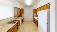 This single-floor unit offers a practical and accessible layout designed with functionality in mind. The floor plan reveals a well-organized arrangement of living spaces, including a spacious main living area, a kitchen, bedrooms, and bathrooms.

The kitchen follows a galley-style configuration, with warm honey-toned wood cabinetry lining both sides of the space. It is equipped with white appliances, including a refrigerator, stovetop, oven, microwave, and dishwasher. Light-toned tile flooring and ample upper and lower cabinetry provide generous storage and prep space.

The living and bedroom areas feature neutral-toned carpet and crisp white walls, creating a clean and bright environment. Windows fitted with horizontal blinds allow natural light to filter in, with views of surrounding greenery. A pendant light fixture adds a functional decorative touch to the main living space.

Both bathrooms are designed with accessibility as a priority, featuring multiple stainless steel grab bars installed near the toilet and bathtub areas. Each bathroom includes a vanity with wood cabinetry, a countertop sink, and light tile flooring. A full bathtub with a shower rod is present in each bathroom.

Overall, the unit presents a straightforward, accessibility-conscious design with consistent neutral finishes throughout.