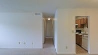 This single-floor residence offers a straightforward and functional layout designed for comfortable everyday living. The interior is finished with a consistently neutral color palette — crisp white walls and trim throughout — creating a clean, bright atmosphere that feels open and airy. Soft wall-to-wall carpeting runs through the main living areas and hallway, adding warmth underfoot and contributing to a quiet, comfortable environment. A central hallway serves as the connective spine of the space, lined with doors that lead to individual rooms, and illuminated by a ceiling-mounted light fixture that ensures the passage remains well-lit. Ceiling-mounted HVAC vents are visible, indicating a centralized climate control system for year-round comfort. Electrical outlets are thoughtfully placed along the walls for practical convenience. To one side, built-in shelving is visible, offering organized storage and display space. The overall design is understated and versatile, providing a neutral canvas well-suited for personalization. The layout flows logically from the main living areas through the hallway to the private rooms, making the space easy to navigate and practical in its arrangement.