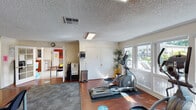 This single-floor fitness and community center is thoughtfully organized to serve multiple purposes. The workout area is well-lit by large windows that frame views of the outdoor surroundings, filling the space with natural light. The fitness zone is equipped with an array of cardio machines, including a treadmill, elliptical trainer, stationary bike, and recumbent bike, all positioned on protective floor mats atop rich hardwood flooring. A water dispenser provides added convenience for users during their workouts.

Accessible through elegant French doors, the adjacent multipurpose room features a warm terracotta-red accent wall adorned with framed artwork, creating a vibrant and welcoming atmosphere. This area accommodates a long conference-style table with multiple chairs, making it suitable for meetings or community gatherings. Additional fitness equipment, including a recumbent bike and wall-mounted television, is also present in this room, adding to its versatility.

The space transitions seamlessly between areas via open doorways and features textured ceilings with flush-mounted fluorescent lighting throughout. The combination of hardwood and carpeted flooring defines each zone clearly, while the overall layout promotes a functional, community-oriented environment ideal for both exercise and collaboration.