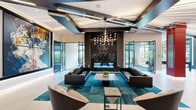 Located at 630 Washington Avenue in Belleville, NJ, this expansive single-floor amenity space delivers a sophisticated, contemporary experience designed for both socializing and productivity. The grand lobby lounge anchors the space with soaring ceilings, bold geometric coffered detailing, and a dramatic sculptural chandelier that casts a warm, ambient glow. Rich leather seating arrangements are set atop vibrant teal area rugs, complemented by a striking floor-to-ceiling black marble feature wall and large-scale abstract artwork that adds an artistic focal point. A built-in bar counter with teal upholstered seating overlooks expansive floor-to-ceiling windows and glass doors, offering direct views of an outdoor courtyard and pool area, flooding the interior with natural light. The games room features a statement red billiard table, large-format display screen, and stylish pendant lighting, ideal for casual entertainment. A dedicated conference room offers a refined setting with a tall marble fireplace surround, an integrated display, soaring angled ceilings, and a generous long-table seating arrangement beneath a modern starburst pendant light. Bold red accent columns and dark exposed beam detailing throughout create a dynamic visual contrast, unifying the space's energetic yet polished design aesthetic.