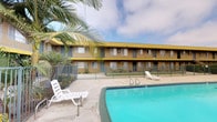 This residential community is centered around a vibrant outdoor amenity area that serves as its social hub. The property features a generously sized swimming pool with clear, blue water, framed by a wide concrete deck that provides ample space for lounging and relaxation. White reclining lounge chairs are positioned poolside, offering a comfortable setting for residents to enjoy the outdoors. A wrought iron security fence encloses the pool area, ensuring a safe and private environment. The surrounding two-story building is finished in a bold yellow exterior with dark trim accents, giving the property a distinctive and easily recognizable character. Units face the courtyard, with windows and individual entry doors lining both levels. A staircase provides access between floors. Mature palm trees and greenery add a touch of tropical landscaping to the setting, complementing the Southern California-inspired aesthetic. The open sky above the courtyard allows for abundant natural light throughout the day, creating a bright and inviting atmosphere. Overall, the property offers a community-oriented layout designed to encourage outdoor living and convenience.