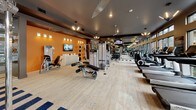 This fitness center is a thoughtfully designed, fully equipped facility that caters to a wide range of workout needs. The space is defined by its striking interior design, featuring a vibrant orange accent wall adorned with wall-mounted lighting fixtures and a flat-screen TV, creating an energetic and motivating atmosphere. The flooring combines light wood-style planks with bold navy and white striped area rugs, adding a dynamic visual element throughout the space.

The strength training area is well-appointed with a Smith machine, cable crossover systems, a variety of weight machines, adjustable benches, and a full rack of free weights, providing ample options for resistance training. Expansive floor-to-ceiling mirrors line one side of the gym, enhancing the sense of openness and allowing users to monitor their form during workouts.

The cardio zone is anchored by a row of modern treadmills positioned directly in front of large windows that frame views of an outdoor courtyard, flooding the area with natural light. Additional cardio equipment, including stationary bikes and ellipticals, rounds out the selection.

Overhead, linear pendant lighting and recessed fixtures illuminate the space with a clean, contemporary feel. The fitness center is accessed through a glass-paneled door, and the overall layout is spacious and well-organized, supporting a comfortable and productive workout experience.