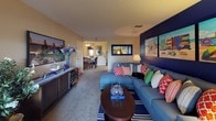 This single-floor residence offers a thoughtfully arranged layout that balances comfort and style. The living area serves as the heart of the home, anchored by a bold navy accent wall that showcases colorful southwestern-inspired artwork, adding a vibrant, regional character to the space. A large sectional sofa dressed in rich blue tones with an array of patterned throw pillows creates a welcoming focal point, complemented by a round dark wood coffee table and a striped area rug beneath. A wall-mounted flat-screen television and a dark wood media console provide both functionality and sleek design on the opposite wall. Soft carpet flooring extends throughout, contributing to the warm and relaxed ambiance. Natural light filters through the interior, enhanced by warm lamp lighting that adds a cozy glow. The living area flows openly into the dining space beyond, which features a ceiling fan and a classic dining table with chairs, creating a seamless transition between entertaining and everyday living. The textured ceiling adds a traditional touch to the overall interior. The space is cohesive, cheerful, and well-suited for comfortable daily living.