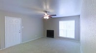 This single-story space centers around a generously sized living area finished with wall-to-wall carpeting in a neutral tone, contributing to a quiet and comfortable ambiance. A fireplace with a dark surround serves as a natural focal point along one wall, adding both visual interest and functional warmth to the room. A ceiling fan with an integrated light fixture is centrally mounted, providing both air circulation and ambient overhead lighting. Natural light enters through a window fitted with horizontal blinds, offering adjustable brightness and privacy. The walls are finished in a soft, neutral beige tone, complementing the overall understated aesthetic. White baseboards run along the perimeter of the room, adding a clean, finished look to the space. The textured ceiling adds subtle architectural detail. The room's open layout and neutral palette create a versatile foundation well-suited to a variety of furnishing arrangements and interior styles.