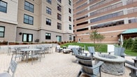 This generously sized outdoor courtyard is designed to offer a well-rounded amenity space for socializing and relaxation. The ground is finished with a herringbone-patterned paver surface throughout, lending a clean and cohesive aesthetic across all zones of the space.

At the heart of the courtyard sits a substantial stone fire pit encircled by Adirondack-style chairs, creating an inviting gathering spot ideal for unwinding. Adjacent to this lounge area, a seating arrangement of upholstered outdoor furniture provides a more intimate setting for conversation.

Multiple dining areas are distributed across the courtyard, each outfitted with modern metal tables and chairs suited for outdoor use. A dedicated outdoor kitchen station, constructed with stacked stone detailing, houses dual stainless steel grills with integrated cabinetry, making it well-equipped for outdoor cooking and entertaining.

Landscaped green lawns and neatly planted trees and shrubs border the paved areas, introducing natural softness to the hardscape surroundings. The courtyard is enclosed by the building's multi-story facades on several sides, offering a sense of shelter while remaining open to natural light and sky above.