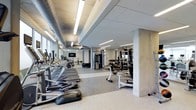 This fitness center offers a comprehensive workout environment designed to accommodate a wide range of exercise needs. The space is laid out across a single floor, with a generous open floor plan that allows for easy navigation between equipment zones. Along one side, a row of cardio machines — including treadmills and ellipticals — lines the perimeter, benefiting from large windows that flood the area with natural light and provide an energizing atmosphere for exercise. The flooring throughout is a durable speckled rubber material, ideal for high-traffic fitness use. Exposed ductwork and linear fluorescent lighting run across the ceiling, giving the space an industrial-modern aesthetic. Structural concrete columns punctuate the layout, adding to the raw, utilitarian character of the design. The strength training area features a cable machine system, a dumbbell rack with a full mirror wall behind it, and a weight bench, making it suitable for a complete resistance training session. A wall-mounted television is also present, providing entertainment or instructional content during workouts. The mirrored walls throughout enhance the sense of spaciousness and allow users to monitor their form. Overall, the fitness center strikes a balance between functionality and modern design.