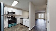 This single-floor unit offers a clean, modern aesthetic with thoughtfully selected finishes throughout. The open-concept kitchen flows seamlessly into the living area, creating a connected and functional layout. The kitchen is well-appointed with white shaker-style cabinetry, granite-look countertops, and a full suite of stainless steel appliances, including a refrigerator, electric range, microwave, and dishwasher. A skylight above the kitchen adds a bright, airy feel to the space. Wood-look plank flooring extends throughout the main living areas, providing both durability and visual warmth.

The living area is generously proportioned and bathed in natural light from a set of French doors that open to the exterior. An additional room, accessible via a windowed door, offers flexible use as a den or office. The bedrooms are finished with plush carpeting and ceiling fans, with windows dressed in blinds to control natural light. The primary bedroom is notably spacious, offering ample room for furnishings.

The bathroom features a white vanity with granite-look countertops, a large mirror with overhead lighting, and wood-look flooring that ties cohesively with the rest of the unit. Neutral gray tones are used throughout the walls, creating a calm, contemporary ambiance that complements the overall design.