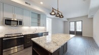 This single-floor residence showcases a thoughtfully designed open-concept layout with contemporary finishes throughout. The kitchen serves as the centerpiece, featuring a striking two-tone cabinetry scheme pairing light gray upper cabinets with dark espresso lower cabinets, all accented with gold hardware. A generously sized granite island with dramatic veining anchors the space beneath a trio of industrial-style brass pendant lights. Stainless steel appliances, including a gas range, over-the-range microwave, refrigerator, and dishwasher, complement the textured white subway tile backsplash. Wood-look flooring flows throughout the kitchen and living areas, which opens to a glass door providing natural light.

The bedroom offers plush carpeting, bright windows with blinds, and access to a spacious walk-in closet outfitted with built-in shelving and dual hanging rods. The full bathroom is well-appointed with a granite-topped white vanity with gold hardware, a tub-shower combination featuring white subway tile surround, stylish wall sconces, and neutral large-format floor tiles. Slate-blue panel doors throughout the unit add a refined, modern touch to the overall aesthetic.