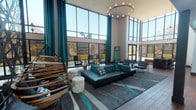 Located at 904 S Mesquite St in Arlington, Texas, this single-floor amenity and leasing space is designed with a bold, contemporary aesthetic that immediately commands attention. The grand lobby entrance features dramatic double-height ceilings framed by floor-to-ceiling windows, flooding the interior with abundant natural light. Teal drapery, rich wood-look flooring, and a statement chandelier set the tone for the vibrant design language found throughout.

The main lounge area is anchored by a large sectional sofa in teal, complemented by eclectic seating arrangements including woven rattan chairs and tufted ottomans, all set atop a geometric area rug. Sleek dark columns provide architectural definition, while a modern fireplace adds warmth to the social atmosphere.

A spacious community kitchen and bar area features a long dark granite island lined with stylish gold-accented bar stools, pendant globe lighting, and wall-mounted flat-screen televisions — ideal for communal gatherings. Adjacent to the lounge, a dedicated leasing office with a dark executive desk and wall-mounted floor plan displays serves as a professional welcome point.

A private conference or study room showcases an eclectic gallery wall with bold pop-art pieces, a large world map, and sculptural décor, paired with a modern dining-style table and contemporary white chairs. Throughout, the space reflects a dynamic, design-forward environment tailored to an engaged, community-oriented lifestyle.