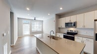 This single-floor residence offers a thoughtfully designed open-concept layout that seamlessly connects the kitchen and living areas. The kitchen is a standout feature, equipped with stainless steel appliances including a side-by-side refrigerator with water dispenser, an electric range, and an over-the-range microwave. Crisp white cabinetry with modern hardware lines the walls, complemented by a classic white subway tile backsplash and warm quartz countertops. A central island with an undermount sink provides additional prep space and a natural gathering point. Rich hardwood-style flooring flows throughout the main living and kitchen areas, adding warmth and continuity to the space. The living area is bathed in natural light through large windows with horizontal blinds and features a ceiling fan, creating a comfortable and airy atmosphere. Recessed lighting throughout the space ensures bright, even illumination. The bedroom is carpeted for comfort and includes a ceiling fan and a generously sized window that invites natural light. The bathroom is finished with a wide quartz vanity, an undermount sink, a large mirror, and direct access to a walk-in closet with open shelving, offering excellent storage and convenience. Neutral gray walls throughout provide a clean, modern backdrop well-suited to a variety of interior styles.