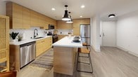 This well-designed single-floor residence offers a cohesive modern aesthetic with warm natural wood tones and clean white finishes throughout. The kitchen is a standout, featuring floor-to-ceiling flat-front cabinetry in a warm oak finish, white quartz countertops, stainless steel appliances including a gas range and built-in microwave, and a geometric tile backsplash. A central island with pendant lighting provides additional prep space and casual seating. The open layout flows into the living area, where wood-look flooring, a neutral color palette, and a glass door that invites natural light from outside create an inviting atmosphere. The primary bedroom offers plush carpeting, a ceiling fan, large windows with blinds and drapery, and ample space for a king-sized setup. A second bedroom mirrors the same comfortable, light-filled character. The primary bathroom includes a sleek walk-in glass shower, a warm wood vanity with white countertop, and a bold round black-framed mirror. The second bathroom features a full soaking tub with shower, wood-toned wall cabinet, and matte black fixtures. Throughout the residence, consistent use of matte black hardware, wood accents, and neutral tones creates a polished, contemporary living environment.