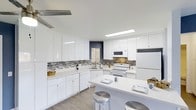 This single-floor space showcases a well-appointed kitchen designed for both functionality and style. The kitchen features an abundance of high-gloss white cabinetry that extends to the ceiling, providing generous storage throughout. A striking mosaic tile backsplash in gray and gold tones adds visual interest and contrasts beautifully against the crisp white surfaces. The layout follows an L-shaped configuration, maximizing workspace efficiency with ample white countertops running along the perimeter. A central island with a clean white surface serves as a casual dining area, accommodated by sleek metallic bar stools. White appliances, including a refrigerator, electric range, and over-the-range microwave, are seamlessly integrated into the design. Light wood-look flooring runs throughout, complementing the overall bright aesthetic. Recessed and flush-mount ceiling lighting ensures the space is well-lit, while a window above the sink allows for natural light. A navy blue accent wall in the background adds a modern, bold touch to the otherwise neutral palette, contributing to the kitchen's contemporary character.