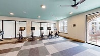 This single-floor fitness studio is thoughtfully designed to support a variety of workout routines in a bright, open environment. The space is divided into two functional zones: a cycling area and a multi-purpose open exercise floor. The cycling section is lined with a row of stationary bikes, each equipped with individual monitor screens, and is backed by large framed mirrors that visually expand the room and allow riders to monitor their form. Recessed lighting and a ceiling fan overhead ensure comfort during intense sessions. The open exercise area offers generous floor space finished with a distinctive patchwork-style carpet in soft neutral tones of grey, beige, and blue, lending the room a modern aesthetic. This zone is equipped with a freestanding dumbbell rack, a rowing machine, yoga mats, and resistance bands, making it versatile for strength training, yoga, or cardio workouts. Colorful artwork decorates the white walls, adding energy and visual interest to the space. Large floor-to-ceiling glass sliding doors along one wall flood the studio with natural light and provide views of an exterior brick-arched courtyard, creating an airy, connected atmosphere. The signature light teal ceiling throughout ties the space together with a fresh, uplifting design touch.