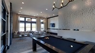 This sophisticated single-floor amenity space seamlessly blends modern design with a welcoming, social atmosphere. At the heart of the room sits a sleek, dark billiards table, positioned beneath a striking branch-inspired chandelier with exposed Edison bulbs that serves as a bold focal point. The open layout flows effortlessly into a comfortable lounge area furnished with plush, neutral-toned sectional sofas and a glass coffee table, creating an inviting space for relaxation and socializing. A wall-mounted flat-screen television is framed by a decorative white laser-cut panel, adding an artistic, textural element to the space. The room is bathed in natural light through expansive floor-to-ceiling windows and glass doors, accented by flowing sheer curtains that soften the contemporary aesthetic. Recessed ceiling lighting supplements the abundant daylight, ensuring the space is well-lit at all hours. Light hardwood flooring and a patterned area rug anchor the lounge area, while clean white walls with subtle dark trim accents contribute to the space's crisp, upscale character. The overall ambiance is polished yet approachable, designed for both lively entertainment and casual gatherings.