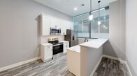 Located at 1230 Elm St in Manchester, NH, this thoughtfully designed single-floor unit offers a comfortable and functional layout across six rooms. The kitchen is a standout feature, boasting white shaker cabinetry, quartz countertops with a subtle veining pattern, a subway tile backsplash, stainless steel appliances including a range, microwave, and refrigerator, and sleek pendant lighting. A peninsula with a matte black faucet adds both workspace and visual appeal, while large floor-to-ceiling windows with blinds flood the area with natural light. Wood-look plank flooring runs through the kitchen and connecting areas, lending warmth and continuity. The carpeted rooms feature neutral gray tones, clean white panel doors with matte black hardware, and recessed lighting, providing flexible spaces suitable for sleeping or other uses. Closet storage with wire shelving is included in one of the carpeted rooms. A bathroom with a white vanity and countertop is conveniently positioned adjacent to an in-unit laundry area equipped with a stacked washer and dryer. Throughout, the space maintains a cohesive, modern aesthetic with light gray walls, crisp white trim, and a neutral palette that creates a clean, move-in-ready environment.