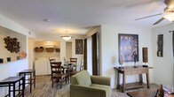 This single-floor residence offers a well-designed, open-concept layout that seamlessly connects the living and dining areas, creating a cohesive and inviting atmosphere throughout. The living area features warm hardwood-style flooring and is furnished with a comfortable sage green armchair, complemented by a rustic wooden console table adorned with decorative artwork. Large framed paintings add a touch of character and visual interest to the neutral, cream-toned walls.

The dining area is elegantly appointed with a round glass-top table surrounded by classic wooden chairs, illuminated by a stylish pendant light that adds a warm glow to the space. Dark curtain panels frame a sliding glass door, allowing natural light to filter in while providing privacy when needed.

Adjacent to the dining space, a laundry area is conveniently integrated into the unit, featuring stacked appliances and built-in shelving with woven baskets for organized storage. The overall ambiance of the space is warm and welcoming, with a thoughtful blend of functional design and decorative touches that create a comfortable, livable environment throughout the single level.