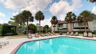 This community amenity space offers a well-rounded array of features designed to enhance everyday living. At its centerpiece is a generously sized outdoor swimming pool with vivid turquoise water, framed by tall palm trees and lush greenery. A row of lounge chairs lines the pool deck, providing ample space for sunbathing, while a shaded seating area with a teal umbrella offers a cool retreat. A playground is visible nearby, adding family-friendly appeal.

Inside, the clubhouse-style space is warm and inviting, with wood-look flooring, soft sage-green walls, and crown molding throughout. The main lounge area features cathedral ceilings with ceiling fans, recessed lighting, and a comfortable black leather seating arrangement centered around a flat-screen television with built-in shelving. Café-style round tables with dark wooden chairs are arranged throughout, encouraging casual gathering.

An entry hallway adorned with framed artwork leads to the upper-level lounge via carpeted steps. The kitchenette is a standout feature, showcasing dark cabinetry with granite countertops, a striking mosaic tile backsplash, pendant lighting, and appliances including microwaves, a toaster oven, and a water dispenser — making this space both functional and stylish.