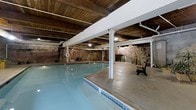 This single-level indoor pool facility offers a distinctive and memorable atmosphere, defined by its remarkable architectural character. The space features exposed natural stone walls that lend a rugged, historic charm, complemented by warm wooden plank ceilings supported by white structural beams and columns throughout. The centerpiece of the space is a generously sized swimming pool with clear blue water, illuminated by underwater lighting that gives the pool a luminous, inviting glow. Blue tile trim lines the pool's edge, adding a decorative touch against the concrete pool surround. The surrounding deck area provides ample space for movement and relaxation, with seating available for guests. Overhead, industrial-style lighting fixtures are mounted to the beams, ensuring the space is well-lit despite its enclosed, subterranean feel. Arched stone passageways are visible along the walls, reinforcing the historic architectural identity of the building and hinting at a storied past. The overall ambiance is one of unique contrast — the rawness of stone and aged wood set against the clean, modern functionality of the pool facility — creating an environment that feels both atmospheric and practical.