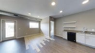This single-floor space showcases a thoughtfully designed open-concept layout that combines living and kitchen areas into one cohesive environment. The flooring features a mix of light and medium-toned hardwood-style planks, creating a visually interesting transition across the space. Neutral greige-toned walls complement the clean white baseboards and trim throughout.

Recessed lighting is installed across the textured ceiling, providing even illumination throughout the main area. A horizontal window along one wall allows natural light to filter in, brightening the interior. An exposed HVAC duct runs along the upper portion of one wall, adding an industrial accent to the otherwise clean aesthetic.

The kitchen area, positioned to one side, includes white lower cabinetry, a dark countertop, and a black appliance, likely a dishwasher. Above, a set of white open shelves provides practical display or storage space. A doorway leads to an adjacent room, visible in the background, which appears to house laundry or utility equipment. Overall, the space offers a clean, modern, and functional design suitable for everyday living.