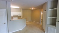 This single-floor unit offers a practical and open layout that seamlessly connects the kitchen and living area. The kitchen is equipped with white cabinetry, a built-in microwave, an electric range, and a peninsula-style breakfast bar that defines the boundary between the cooking and living spaces. The living area is finished with plush wall-to-wall carpeting in a neutral tone, creating a warm and comfortable environment. Walls are painted in a soft beige, contributing to a cohesive and understated aesthetic throughout. Overhead flush-mount lighting illuminates the interior, complemented by a fluorescent fixture above the kitchen workspace. A hallway visible toward the rear of the unit leads to additional rooms, suggesting a functional and well-organized floor plan. A closet with classic louvered bifold doors is conveniently positioned near the living area, providing accessible storage. Clean white baseboards trim the space throughout, adding a crisp finishing detail. Overall, the unit presents a straightforward, move-in-ready layout well-suited for everyday living.