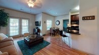 This single-floor residence offers a thoughtfully designed open-concept layout that seamlessly connects the living, dining, and kitchen areas into one cohesive and inviting space. Warm hardwood flooring flows throughout the main living areas, complemented by neutral-toned walls adorned with crown molding that adds a refined architectural touch.

The living room is generously proportioned, featuring a ceiling fan with light fixture, large windows with blinds that invite abundant natural light, and French doors that open to an outdoor area. The space accommodates comfortable seating and offers a welcoming ambiance.

Adjacent to the living room, the dining area provides ample room for a full dining set, positioned near windows that keep the space bright. The kitchen is semi-open, separated by a breakfast bar with dark granite countertops and bar seating. Rich dark wood cabinetry, black appliances, and a teal accent wall give the kitchen a bold yet polished character.

A dedicated entryway greets visitors with a solid panel front door equipped with a secure deadbolt and lever handle, alongside a side table with warm lamp lighting. The overall design balances practicality with style, creating a comfortable and functional living environment with consistent finishes and a cohesive color palette throughout.