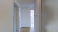 This single-floor space is characterized by its clean, minimal aesthetic and bright interior. The hallway serves as the central connective element of the layout, finished in crisp white walls that reflect natural light streaming in from windows visible at the far end. The flooring throughout the corridor consists of light-toned carpet, contributing a soft, understated feel to the space. A flush ceiling fixture provides supplemental overhead lighting, while a thermostat mounted on the wall indicates climate control functionality. Doorways branch off from the hallway on either side, offering access to adjoining rooms. The open doorway at the end of the corridor leads to a well-lit room with a window, suggesting good natural light penetration throughout. The overall ambiance is understated and functional, with a neutral color palette of whites and beiges creating a cohesive, airy atmosphere. The straightforward layout and uncluttered design lend the space a sense of practicality, making it well-suited for a variety of uses. The single-floor configuration ensures easy accessibility and a seamless flow between areas.
