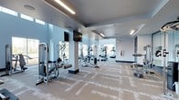 This fitness center offers a thoughtfully designed, single-floor layout that provides ample space for a comprehensive workout experience. The open floor plan is finished with patterned, neutral-toned carpet tiles that lend a clean, contemporary aesthetic throughout the space.

The facility is outfitted with a variety of strength training machines and cable systems positioned along the perimeter, complemented by a dedicated cardio section featuring treadmills and elliptical machines. Wall-mounted flat-screen televisions are strategically placed to keep members engaged during workouts. A free-weight station with a bench is also available for additional training versatility.

Natural light flows in through large windows along the far wall, creating a bright and energizing atmosphere. The space is further illuminated by recessed linear LED lighting installed in a tray ceiling, which adds a modern architectural touch while ensuring consistent, even lighting across the entire floor.

The walls are finished in a crisp, light blue-gray palette that contributes to the clean and refreshing ambiance of the space. Structural columns are accented in a deeper charcoal tone, adding visual contrast and dimension. Overall, this fitness center combines functional equipment with a polished, modern design to deliver an inviting and motivating environment for users of all fitness levels.