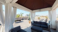 Located in the heart of Hollywood at 5711 Hollywood Blvd, this rooftop terrace offers an expansive open-air retreat with sweeping panoramic views of the Los Angeles skyline, palm tree-lined streets, and the surrounding Hollywood Hills. The space is anchored by a stylish octagonal cabana structure featuring a warm wood-toned ceiling and flowing white drape curtains that can be drawn for shade or privacy. Inside the cabana, comfortable dark navy upholstered lounge seating and an ottoman create an inviting gathering area, while an adjacent outdoor dining table with seating accommodates group meals or meetings. Beyond the cabana, the open terrace provides dedicated zones for outdoor entertaining, including multiple stainless steel gas grills and a wicker dining table — ideal for al fresco cooking and dining. A dedicated sun-bathing area features two reclining chaise lounges surrounded by lush potted palms, succulents, and tropical plantings that lend a resort-like ambiance. A balcony ledge on the perimeter offers additional vantage points over the Hollywood streetscape below. The terrace receives abundant natural sunlight throughout the day, making it a versatile space suited for relaxation, social gatherings, or outdoor events in one of Los Angeles's most iconic neighborhoods.