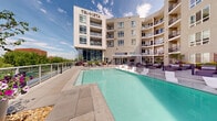 Located at 1350 Speer Blvd in Denver, CO, the Speer Social & Pool and Spa Deck at Tri·Vista is a thoughtfully designed amenity space that seamlessly blends indoor comfort with outdoor recreation. The outdoor deck showcases a large, resort-style swimming pool with crystal-clear turquoise water, complemented by a separate spa. The pool deck is lined with sleek lounge chairs and decorative planters filled with vibrant flowers, all set against a backdrop of the Tri·Vista building's contemporary multi-story façade. Glass railings along the perimeter open up views of the surrounding Denver cityscape and lush greenery. A covered pergola-style transition area with floor-to-ceiling glass doors connects the outdoor deck to the interior social lounge, allowing abundant natural light to flow through. Inside, the social lounge features soaring ceilings, large-format tile flooring, and a striking floor-to-ceiling mosaic tile accent wall housing a fireplace and flat-screen television. A stylish bar counter with white quartz surfaces and upholstered bar seating anchors the space, backed by illuminated shelving and a detailed tile backsplash. Amber pendant lighting adds warmth, while a wet bar area with a brushed gold faucet and stone countertop provides functional entertaining amenities. The overall ambiance is modern, upscale, and inviting.