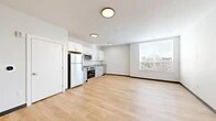 Located at 222 Canal St in New Haven, CT, this single-floor unit offers a clean, modern design with a highly functional open layout. The combined kitchen and living area forms the heart of the space, bathed in natural light from a large window that frames views of the surrounding New Haven cityscape. Light hardwood flooring runs continuously throughout, lending warmth and visual continuity to the interior.

The kitchen is well-appointed with crisp white shaker-style cabinetry, granite countertops, and a full suite of stainless steel appliances, including a refrigerator, range, over-the-range microwave, and dishwasher. Open shelving accents the upper cabinetry, adding a contemporary touch. Flush-mount ceiling fixtures provide consistent ambient lighting across the open living area, which offers a generous, flexible space well-suited for various furniture arrangements.

The bathroom is finished in a neutral, modern palette featuring large-format tile flooring, a walk-in shower with built-in corner shelving, a vanity with a white quartz countertop and undermount sink, and a wide mirror with overhead lighting. Throughout the unit, the clean white walls and minimalist finishes create a bright, cohesive ambiance that reflects a contemporary urban aesthetic.