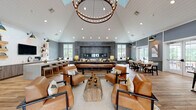 Located at 500 Barclay Blvd in Marlton, NJ, this single-story clubhouse is a thoughtfully designed community amenity space that blends modern rustic aesthetics with upscale comfort. The centerpiece of the space is a dramatic vaulted ceiling clad in white shiplap, anchored by a striking tiered circular chandelier that draws the eye upward and fills the room with warm, ambient light. A bold dark-tiled accent wall serves as a dramatic focal point, flanked by large windows that invite abundant natural light throughout the interior.

The main lounge area features rich cognac leather armchairs arranged around a live-edge wood coffee table, creating an inviting setting for casual gatherings. A full-length shuffleboard table occupies the middle of the room, adding a recreational element to the social space. A built-in banquette and dark upholstered sofas provide additional seating alongside cowhide-accented rugs that reinforce the space's contemporary lodge character.

A well-appointed café-style bar area includes a quartz countertop, wood-toned cabinetry, a coffee station, beverage refrigerator, and reclaimed wood floating shelves. Smaller bistro and dining tables offer intimate seating nooks. French doors throughout open directly to an outdoor patio, seamlessly connecting the interior to the surrounding outdoor amenities. Wood-look tile flooring ties the entire space together with warmth and cohesion.