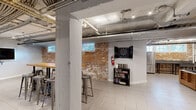 Located at 200 Rhode Island Ave NE in Washington, DC, this single-floor amenity space delivers a compelling industrial-chic aesthetic across two distinct areas. The design draws character from exposed brick accent walls, open ceilings with visible ductwork and conduit, and light-toned tile flooring that unifies the layout throughout.

The main lounge area is anchored by a generously sized sectional sofa with a wood-frame base surrounding a glass-top coffee table, creating a comfortable gathering hub. A wall-mounted flat-screen TV, foosball table, and additional seating arrangements make this zone ideal for socializing and entertainment. Recessed track lighting illuminates the space, while small windows along the brick walls allow in touches of natural light.

The second area serves as a dedicated game and activity room, featuring a full-length shuffleboard table paired with another wall-mounted TV, and a built-in bar-height counter with metal stools for spectators. A dartboard cabinet and a bookshelf with games and books add further recreational options.

A functional kitchenette area includes a stainless steel refrigerator and an industrial-style utility sink, supporting events and gatherings. Throughout the space, the raw architectural elements — from the exposed overhead systems to the weathered brick — combine with modern furnishings to create an urban, community-focused environment well-suited for residents or tenants seeking shared amenity space.