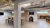 Located at 200 Rhode Island Ave NE in Washington, DC, this single-floor amenity space delivers a compelling industrial-chic aesthetic across two distinct areas. The design draws character from exposed brick accent walls, open ceilings with visible ductwork and conduit, and light-toned tile flooring that unifies the layout throughout.

The main lounge area is anchored by a generously sized sectional sofa with a wood-frame base surrounding a glass-top coffee table, creating a comfortable gathering hub. A wall-mounted flat-screen TV, foosball table, and additional seating arrangements make this zone ideal for socializing and entertainment. Recessed track lighting illuminates the space, while small windows along the brick walls allow in touches of natural light.

The second area serves as a dedicated game and activity room, featuring a full-length shuffleboard table paired with another wall-mounted TV, and a built-in bar-height counter with metal stools for spectators. A dartboard cabinet and a bookshelf with games and books add further recreational options.

A functional kitchenette area includes a stainless steel refrigerator and an industrial-style utility sink, supporting events and gatherings. Throughout the space, the raw architectural elements — from the exposed overhead systems to the weathered brick — combine with modern furnishings to create an urban, community-focused environment well-suited for residents or tenants seeking shared amenity space.