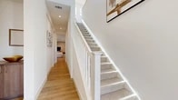Located at 47432 Iris Way in La Quinta, California, this two-story home showcases a clean, contemporary design with a cohesive palette of neutral tones and modern finishes throughout. The entry level features warm wood-look flooring that flows through the main living areas, complemented by crisp white walls and recessed lighting. A staircase with a white-painted banister and carpeted treads connects the two floors, with natural light filtering in from upper windows to brighten the corridor.

The kitchen is outfitted with dual-tone cabinetry — white uppers paired with warm wood lower cabinets — white quartz countertops, a stainless steel appliance suite, and a breakfast bar for casual dining. Adjacent spaces provide additional room for dining or transitional use. The living area is bright and welcoming, featuring large windows with roller shades that bring in natural light.

A discreet water closet on the main level offers added convenience. Upstairs, the bedrooms are carpeted and comfortably scaled, with closet storage included. Bathrooms feature white tile flooring, wood-toned vanity cabinetry, white countertops, and glass-enclosed showers. A carpeted storage or utility space completes the upper level.
