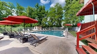 Located in West Chester, PA, this outdoor pool facility offers a well-appointed recreational retreat designed for relaxation and leisure. The centerpiece is a generously sized swimming pool with clear, blue water, surrounded by a wide concrete sundeck that provides ample space for lounging and socializing. The deck is furnished with multiple sling-style lounge chairs and bold red patio umbrellas that offer shaded seating throughout the day, creating a vibrant and inviting atmosphere. A raised wooden deck area with red awning accents is positioned adjacent to the main building, providing an additional gathering space that connects seamlessly to the pool area. A small utility shed with a red-trimmed door sits along the perimeter, offering convenient on-site storage for pool equipment. The entire space is enclosed by a white metal fence, ensuring both security and a clean, finished aesthetic. Lush, mature trees surround the facility on all sides, providing a natural canopy that adds privacy and a sense of seclusion from the surrounding area. A neatly maintained grass lawn borders part of the pool deck, with white Adirondack-style chairs positioned for quiet relaxation under the shade of the trees. The overall ambiance is bright, open, and refreshing, making this pool area an ideal outdoor amenity.