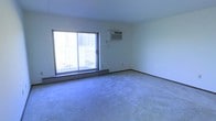 This single-floor space features a spacious, open living area finished with wall-to-wall blue-grey carpeting that adds a soft, neutral tone throughout. The room is kept bright by a large sliding glass door that opens to a private balcony, allowing natural light to fill the interior while offering a direct outdoor access point. A baseboard heater runs along the exterior wall beneath the window, providing efficient in-room heating. Climate control is further supported by a wall-mounted air conditioning unit positioned above the sliding door, ensuring year-round comfort. The walls are clean and white, offering a blank canvas adaptable to a variety of interior styles. The overall layout is open and unobstructed, lending a sense of generous square footage that can accommodate flexible furniture arrangements. The space presents a functional and straightforward design, well-suited for comfortable everyday living.