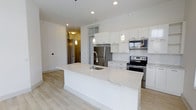 This single-floor unit showcases a bright, open-concept kitchen and living area designed with clean, modern aesthetics. The kitchen serves as the focal point, anchored by a generously sized island with a white quartz countertop, an undermount sink, and a pull-down faucet — providing ample workspace and a natural gathering spot. White flat-panel cabinetry lines the walls, complemented by a classic white subway tile backsplash that adds texture while maintaining a cohesive palette. Stainless steel appliances, including a French door refrigerator, electric range, and built-in microwave, deliver a polished, contemporary look. Pendant lighting fixtures hang above the island, contributing warm, focused illumination alongside recessed ceiling lights. Light-toned engineered wood flooring runs throughout, enhancing the sense of openness and continuity. The white walls and ceiling amplify natural and artificial light, creating a bright and airy atmosphere. A hallway visible from the kitchen leads toward the entryway, suggesting a thoughtfully laid-out floor plan. Overall, the space offers a fresh, minimalist environment suited for modern living.
