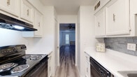This single-story residence presents a clean, neutral aesthetic throughout its well-organized layout. The galley-style kitchen is a functional highlight, featuring white raised-panel cabinetry with brushed nickel hardware, white countertops, a gray tile backsplash, and black appliances including an electric range, dishwasher, and refrigerator. Light wood-look laminate flooring runs through the kitchen, creating a cohesive and easy-to-maintain surface.

The spacious living area benefits from multiple windows fitted with horizontal blinds, allowing natural light to filter in while maintaining privacy. The textured ceiling adds dimension to the open feel of the room. Wood-look laminate flooring continues here, tying the main living spaces together seamlessly.

The bedroom features plush carpeting, a ceiling fan with light fixture, a louvered closet door, and built-in open shelving, offering practical storage solutions. A second bedroom includes a sliding closet with a shelf and hanging rod. Both rooms maintain the space's consistent bright, neutral palette.

The bathroom is finished in crisp white tones, with a vanity featuring ample cabinet storage, a mirror, and a separate shower area with a window for natural light. Overall, the space offers a well-maintained, functional design with consistent finishes throughout.