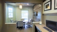 This single-floor office space offers a polished, professional environment thoughtfully designed with a blend of modern and rustic aesthetics. The focal point is a well-appointed conference or meeting room, partially enclosed by glass partitions that maintain an open, connected feel while providing a defined workspace. Inside, a dark conference table is surrounded by upholstered rolling chairs, creating a comfortable setting for meetings or collaborative work.

A striking reclaimed wood accent wall adds warmth and texture, contrasting beautifully with the soft sage-green painted walls. A wall-mounted flat-screen display is positioned on the wood feature wall, adding functional versatility to the room. Natural light filters in through a window dressed with a patterned fabric valance and a neutral roller shade, brightening the interior. An elegant geometric pendant light fixture hangs overhead, providing both ambient illumination and a decorative focal point.

Adjacent to the conference room, a workstation or reception counter is visible, suggesting a multi-functional layout suited for both client-facing interactions and administrative tasks. The overall ambiance is inviting, organized, and professionally styled.