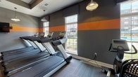 This modern fitness center is thoughtfully designed to support a full range of workout routines across a single open floor. The space is unified by a striking gray and orange color palette, with a bold horizontal orange stripe running along the walls and complementary orange accent tiles set into the patterned carpet flooring, creating a cohesive and energizing aesthetic throughout.

The main workout area is generously sized and features a comprehensive selection of strength training machines, including cable systems, a multi-function weight machine, and adjustable benches, along with a dedicated free weights section equipped with a full dumbbell rack. A row of treadmills lines one wall, accompanied by elliptical trainers and other cardio equipment, all positioned near large windows that invite natural light and provide views of the surrounding outdoor environment.

A separate functional training zone is stocked with stability balls, medicine balls, resistance equipment, yoga mats, and a wall-mounted flat-screen television for guided workouts. Additional wall-mounted TVs are present in the main area as well, ensuring entertainment and instructional content is accessible throughout the space.

Pendant lighting fixtures hang from the tray ceilings, adding an industrial-modern touch that complements the overall design. The fitness center is well-ventilated and organized to accommodate multiple users simultaneously, making it a practical and motivating environment for all fitness levels.