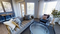 This single-floor residence offers a thoughtfully designed living space that balances comfort and contemporary style. The living room serves as the centerpiece of the home, bathed in abundant natural light streaming through multiple large windows dressed with elegant white drapes and adjustable blinds. The generous window arrangement provides pleasant views of the surrounding greenery, creating a seamless connection between indoors and outdoors.

A glass-panel door leads directly to a private balcony, further enhancing the sense of openness and allowing for easy outdoor access. The living area is anchored by a tufted, gray upholstered sofa accented with layered throw pillows in neutral tones, paired with a sleek round glass coffee table that adds a modern, airy touch. A dark metal accent chair with a blush cushion complements the refined color palette of soft grays, whites, and warm neutrals.

The flooring features rich hardwood-style planks that add warmth and visual continuity throughout the space. A wall-mounted unit and strategically placed furniture ensure both functionality and flow. The overall ambiance is calm, bright, and welcoming, making this a comfortable and stylish living environment suited for everyday relaxation and entertaining alike.