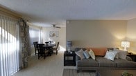 This single-floor residence showcases an open-concept layout that seamlessly connects the living and dining areas into one cohesive, functional space. The living area is anchored by a generously sized sofa adorned with decorative throw pillows in teal and neutral tones, complemented by a dark-finished coffee table that adds contrast to the light-toned carpet flooring running throughout. A patterned area rug defines the seating zone, adding texture and visual interest to the room.

Adjacent to the living space, the dining area accommodates a dark wood round table paired with matching chairs, positioned near a large window dressed with vertical blinds that allow natural light to filter softly into the room. A ceiling fan with a light fixture is installed above the dining area, offering both ventilation and overhead illumination.

The walls are finished in a warm neutral tone, providing a versatile backdrop throughout the space. A textured popcorn ceiling adds a classic character overhead. Decorative touches, including a table lamp with a geometric-patterned shade and floral wall art, contribute to the approachable, comfortable ambiance of this well-organized home.