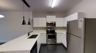 This two-floor space showcases a thoughtfully designed interior with a strong emphasis on modern aesthetics. The kitchen is a standout feature, characterized by clean-lined white cabinetry that extends to the ceiling, maximizing storage while maintaining a streamlined appearance. Crisp white countertops provide ample workspace and contrast elegantly against the dark charcoal tile flooring, creating a bold visual balance.

The kitchen is fully equipped with a complete suite of stainless steel appliances, including a freestanding electric range with oven, an over-the-range microwave, and a full-sized refrigerator. A deep undermount sink with a matte black faucet is integrated into the peninsula-style counter, which also serves as a natural divider between the kitchen and adjoining living areas. Three black pendant lights hang above the peninsula, adding a contemporary design element while providing focused task lighting.

The overall color palette — neutral grays, crisp whites, and matte black accents — lends the space a cohesive, modern character. Recessed and overhead fluorescent lighting ensures the kitchen remains bright and functional throughout the day.